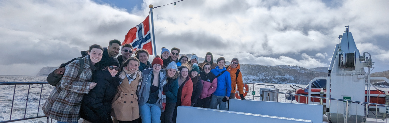 A group of students stands in warm clothes against a railing separating them from the wavy waters behind. They stand in front of a Norwegian flag, and there are many snow-covered mountains in the back. 
